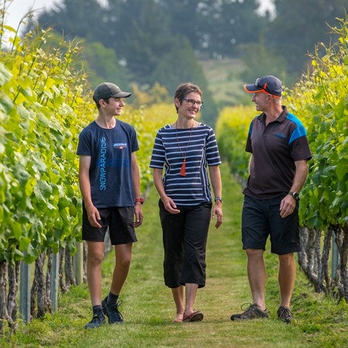 Owners Daniel and Ursula Schwarzenbach with son Thomas walking through their vineyard while chatting. The leaves are bright green and in the background there are some pine trees.
