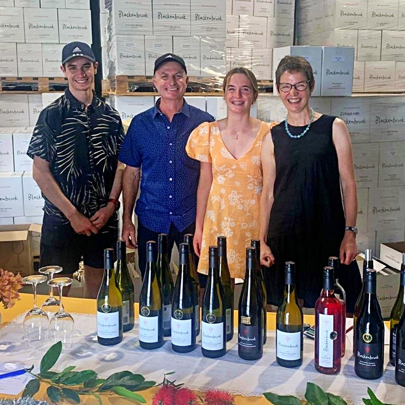 The Schwarzenbach Family (from left: Thomas, Daniel, Isabelle and Ursula) during a wine tasting at their winery.  They stand behind a table with Blackenbrook bottles, some glasses and flowers for decoration.  In the background you can see the Blackenbrook cartons neatly stacked onto pallets. 