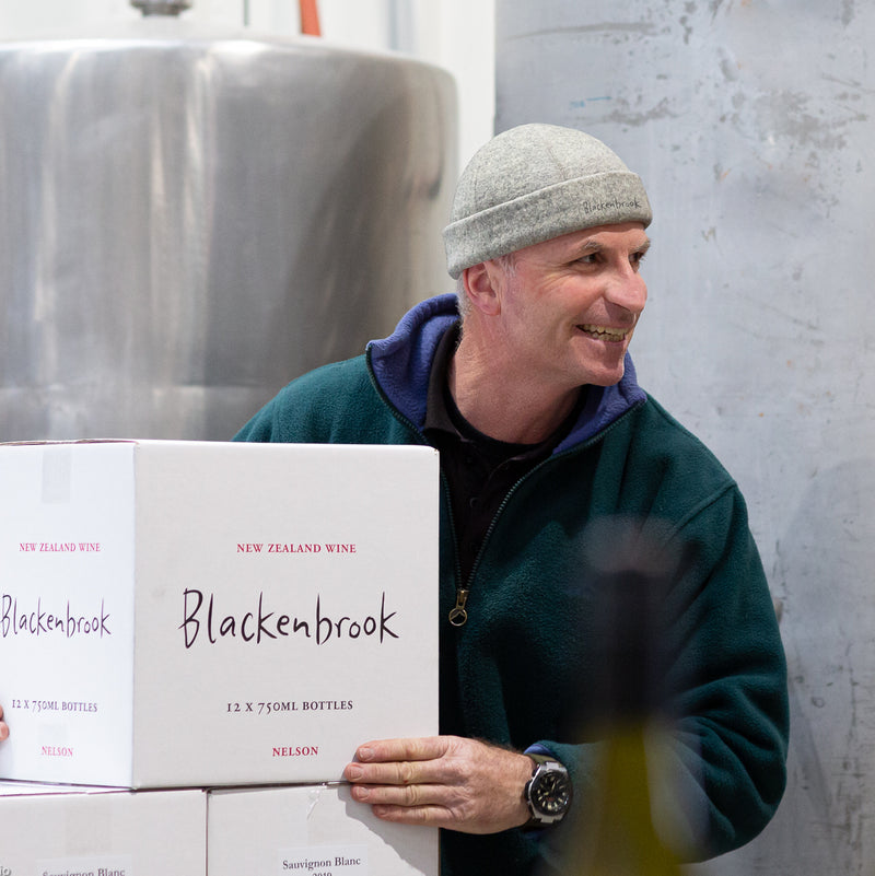Winemaker Daniel Schwarzenbach carrying a case of Blackenbrook wine through the winery.  He is wearing a dark green jersey and a grey hat. In the background there are some stainless steel wine tanks.  