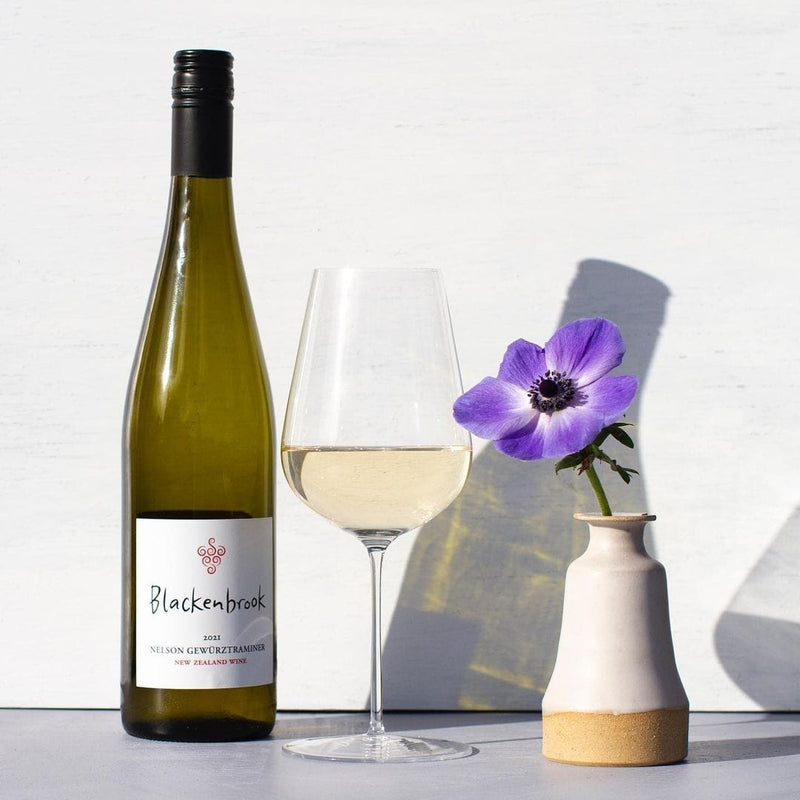 A bottle of Blackenbrook Gewurztraminer next to a glass half full of white wine and a white vase with one purple flower standing on a white surface.  The background is white, too. 