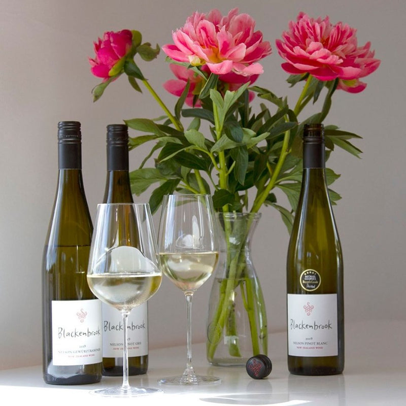Three bottles of Blackenbrook wine with two half-full glasses and a vase with pink peonies on a white table.
