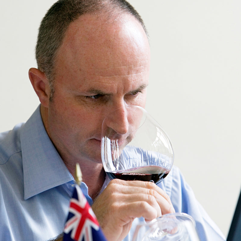 Daniel Schwarzenbach acting as a wine judge at the Mundus Vini International Wine Competition in Germany.  He has a glass of red wine in his hand.  In front of him is the New Zealand flag.