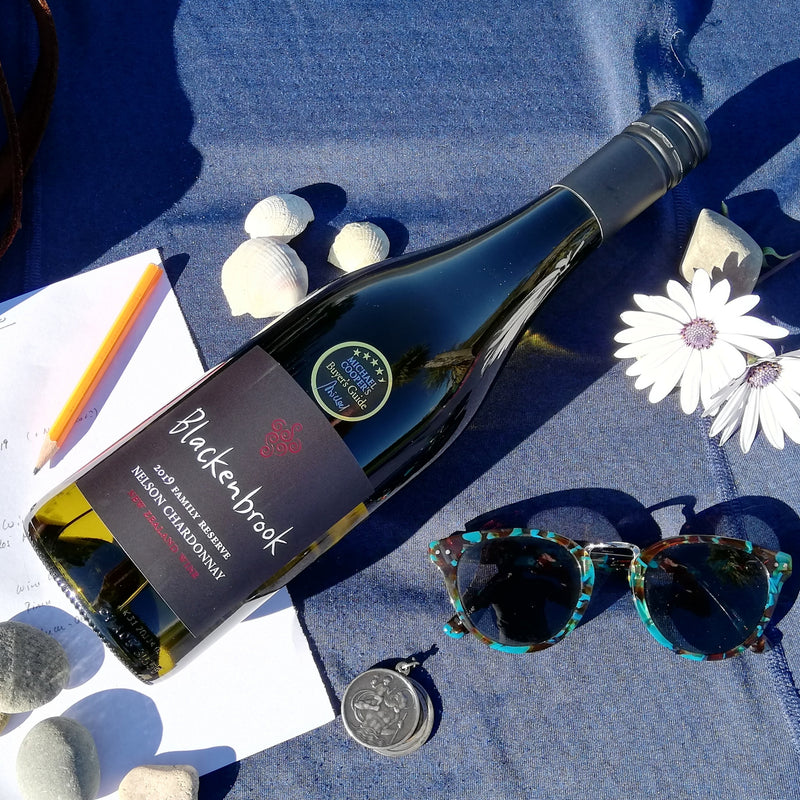 A bottle of Blackenbrook Family Reserve Chardonnay 2019 laying on a blue table cloth.  Around the bottle there are white flowers, mussel shells, pebbles, a pencil and a pair of sunglasses - a very summery and relaxed image.