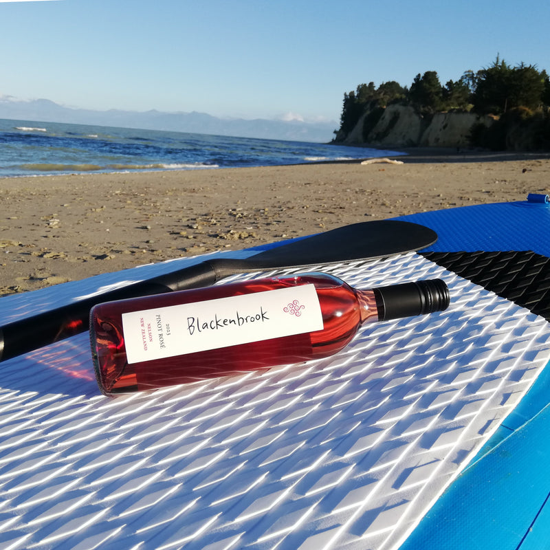 A bottle of dark pink Blackenbrook Rose resting on the white, textured surface of a paddle board.  In the background you can see the beach, some trees, the sea and the Richmond Ranges.