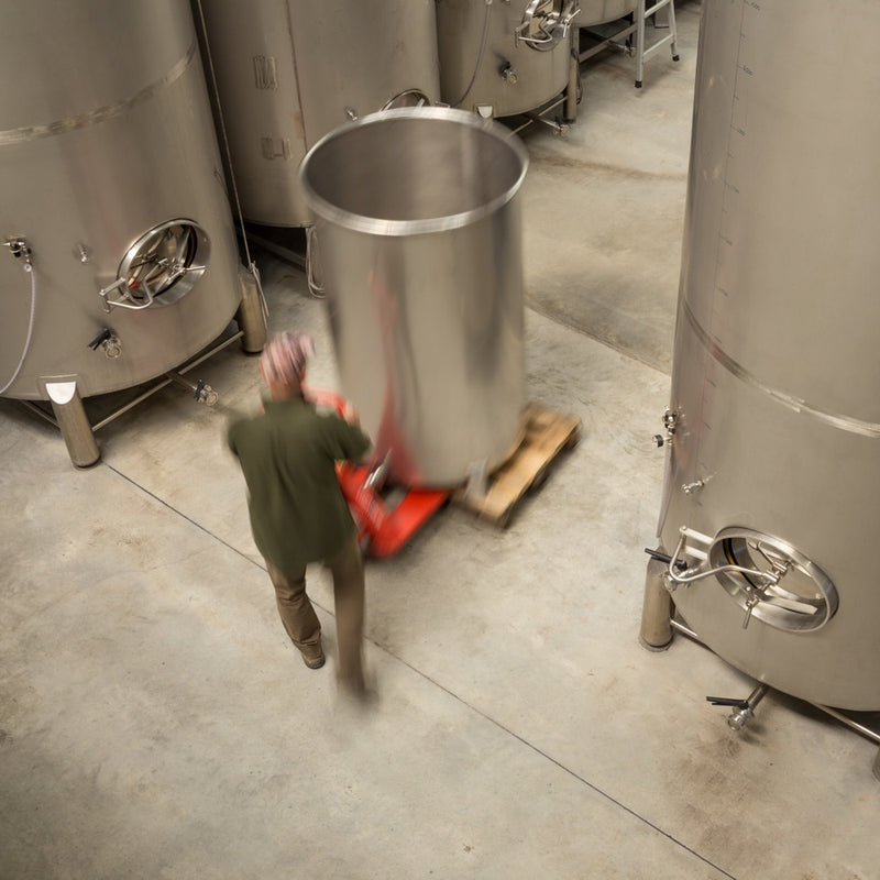 View from the Winery's top floor down into the main winery. Winemaker Daniel Schwarzenbach moves a small stainless steel tank around with a red pallet trolley.  On both sides there are large wine tanks and the floor is concrete.    