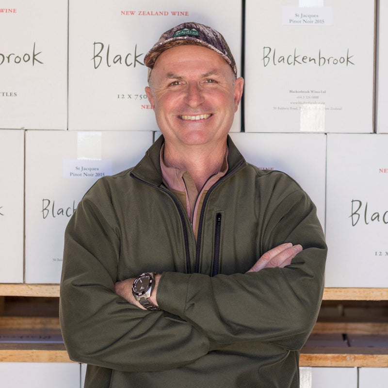 Winemaker Daniel Schwarzenbach standing in front of white Blackenbrook wine boxes.  He is smiling, wearing an olive green jersey and a cap.   