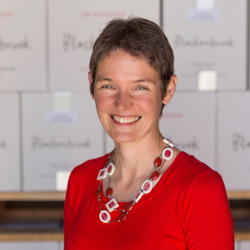 Owner and Marketing Manager Ursula Schwarzenbach standing in front of a pallet of Blackenbrook wine cartons.  She is smiling, wearing a bright red top and beautiful necklace.