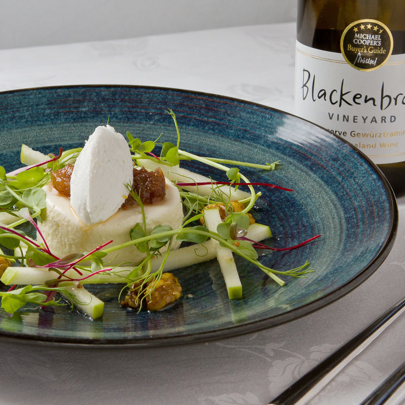 A blue plate with vegan semolina mousse on a bed of pear and water cress salad.  In the background there is a bottle of Blackenbrook Reserve Gewurztraminer with a 4 1/2 Stars Michael Cooper award sticker.