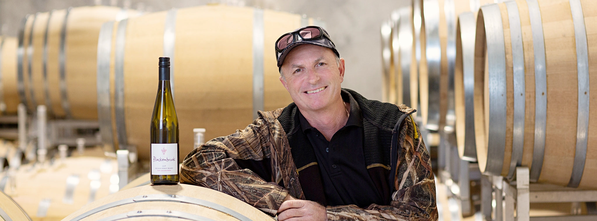 Daniel Schwarzenbach in the barrel room of the Blackenbrook Winery.  There is a bottle of Pinot Blanc standing on a barrique and in the background you can see stacks of full barrels. 